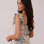 A woman is standing against a white backdrop facing the back and looking over her shoulder. She's wearing a tea-length cream dress with a delicate blue floral print. The dress has a ruffle detail around the neckline that can be worn on or off the shoulder. The skirt has a slit up to the thigh and a ruffle detail around the hem. This is the Renfrew Dress in Ayla. 