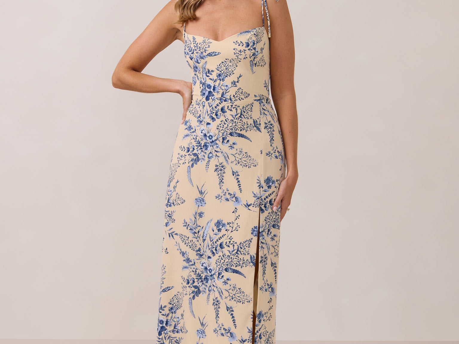 A woman is standing against a white backdrop wearing a cream colored dress with a delicate blue floral print. The dress has tie spaghetti straps and a slit up the skirt. This is the Willow Dress in Ayla. 