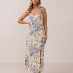 A woman is standing against a white backdrop wearing a cream colored dress with a delicate blue floral print. The dress has tie spaghetti straps and a slit up the skirt. This is the Willow Dress in Ayla. 