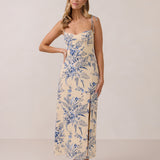 A woman is standing against a white backdrop wearing a cream colored dress with a delicate blue floral print. The dress has tie spaghetti straps and a slit up the skirt. This is the Willow Dress in Ayla. 