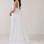 A woman standing in front of a white backdrop is wearing a simple white wedding dress with a long, floor length chiffon neck scarf that trails beautifully on the ground. This is the Maeve bridal scarf.