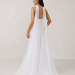 A woman standing facing the back in front of a white backdrop is wearing a simple white wedding dress with a long, floor length chiffon neck scarf that trails beautifully on the ground. This is the Maeve bridal scarf.