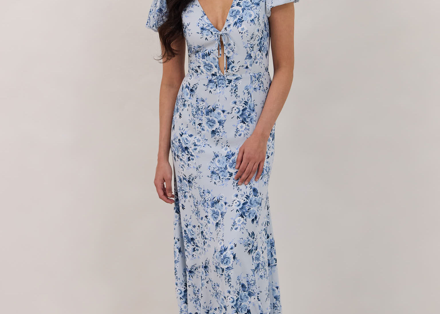 A woman is wearing the Beatrice Dress in Lila. This is a light blue dress with a beautiful blue floral print. The dress has a slightly puffed sleeve, and a full back with a keyhole detail. The front has a deep-V cut with a delicate tie at the bust and another keyhole detail down to the waistline. This is a bra-friendly dress that is ankle length. 