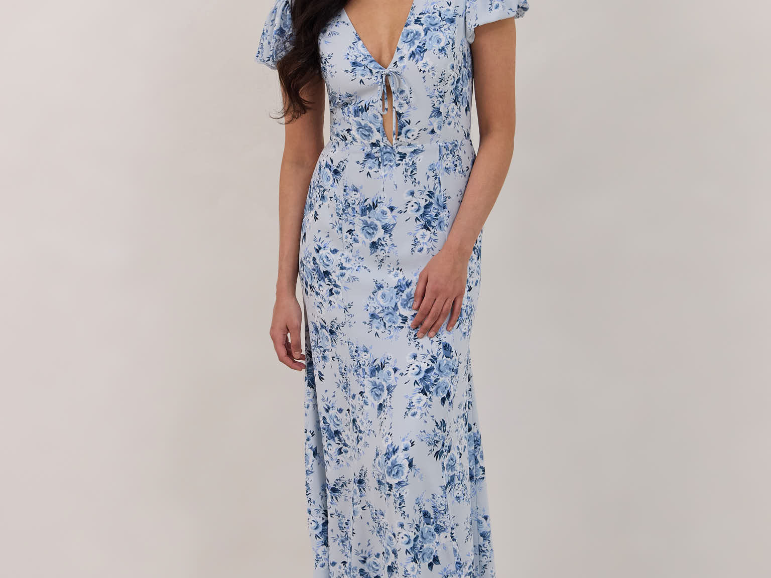 A woman is wearing the Beatrice Dress in Lila. This is a light blue dress with a beautiful blue floral print. The dress has a slightly puffed sleeve, and a full back with a keyhole detail. The front has a deep-V cut with a delicate tie at the bust and another keyhole detail down to the waistline. This is a bra-friendly dress that is ankle length. 
