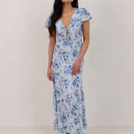 A woman is wearing the Beatrice Dress in Lila. This is a light blue dress with a beautiful blue floral print. The dress has a slightly puffed sleeve, and a full back with a keyhole detail. The front has a deep-V cut with a delicate tie at the bust and another keyhole detail down to the waistline. This is a bra-friendly dress that is ankle length. 