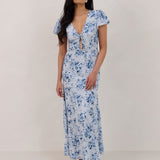 A woman is wearing the Beatrice Dress in Lila. This is a light blue dress with a beautiful blue floral print. The dress has a slightly puffed sleeve, and a full back with a keyhole detail. The front has a deep-V cut with a delicate tie at the bust and another keyhole detail down to the waistline. This is a bra-friendly dress that is ankle length. 