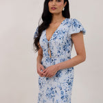 A woman is wearing the Beatrice Dress in Lila. This is a light blue dress with a beautiful blue floral print. The dress has a slightly puffed sleeve, and a full back with a keyhole detail. The front has a deep-V cut with a delicate tie at the bust and another keyhole detail down to the waistline. This is a bra-friendly dress that is ankle length. 