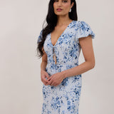 A woman is wearing the Beatrice Dress in Lila. This is a light blue dress with a beautiful blue floral print. The dress has a slightly puffed sleeve, and a full back with a keyhole detail. The front has a deep-V cut with a delicate tie at the bust and another keyhole detail down to the waistline. This is a bra-friendly dress that is ankle length. 
