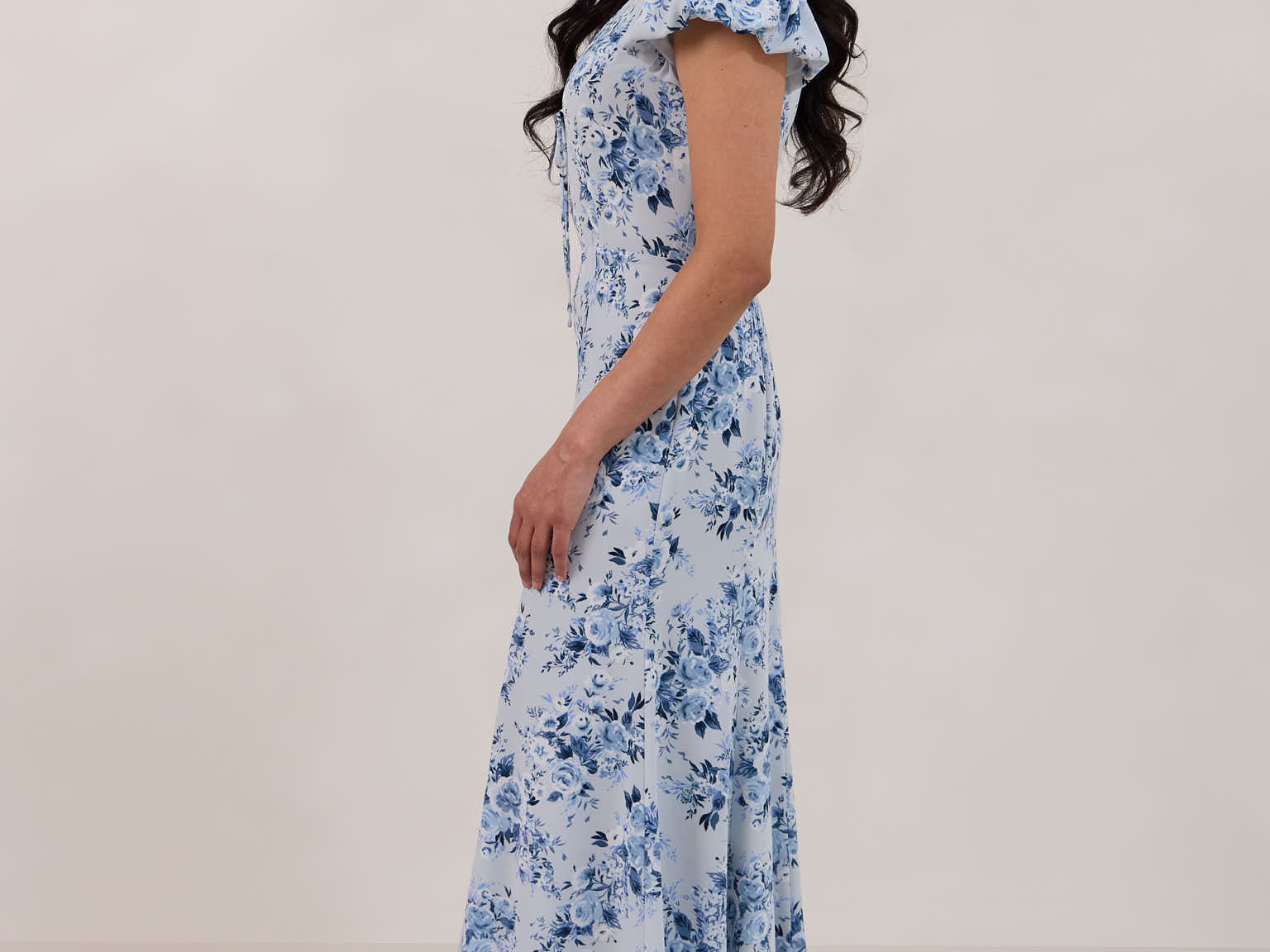A woman is wearing the Beatrice Dress in Lila. This is a light blue dress with a beautiful blue floral print. The dress has a slightly puffed sleeve, and a full back with a keyhole detail. This is a bra-friendly dress that is ankle length. 