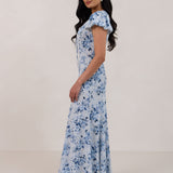 A woman is wearing the Beatrice Dress in Lila. This is a light blue dress with a beautiful blue floral print. The dress has a slightly puffed sleeve, and a full back with a keyhole detail. This is a bra-friendly dress that is ankle length. 
