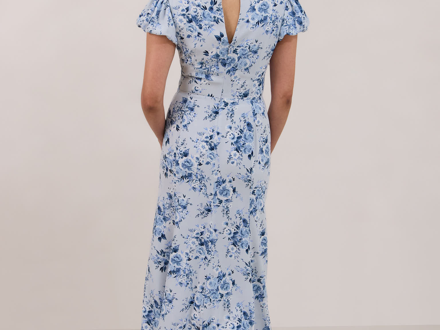 A woman is wearing the Beatrice Dress in Lila. This is a light blue dress with a beautiful blue floral print. The dress has a slightly puffed sleeve, and a full back with a keyhole detail. This is a bra-friendly dress that is ankle length. 