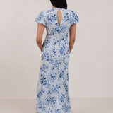 A woman is wearing the Beatrice Dress in Lila. This is a light blue dress with a beautiful blue floral print. The dress has a slightly puffed sleeve, and a full back with a keyhole detail. This is a bra-friendly dress that is ankle length. 