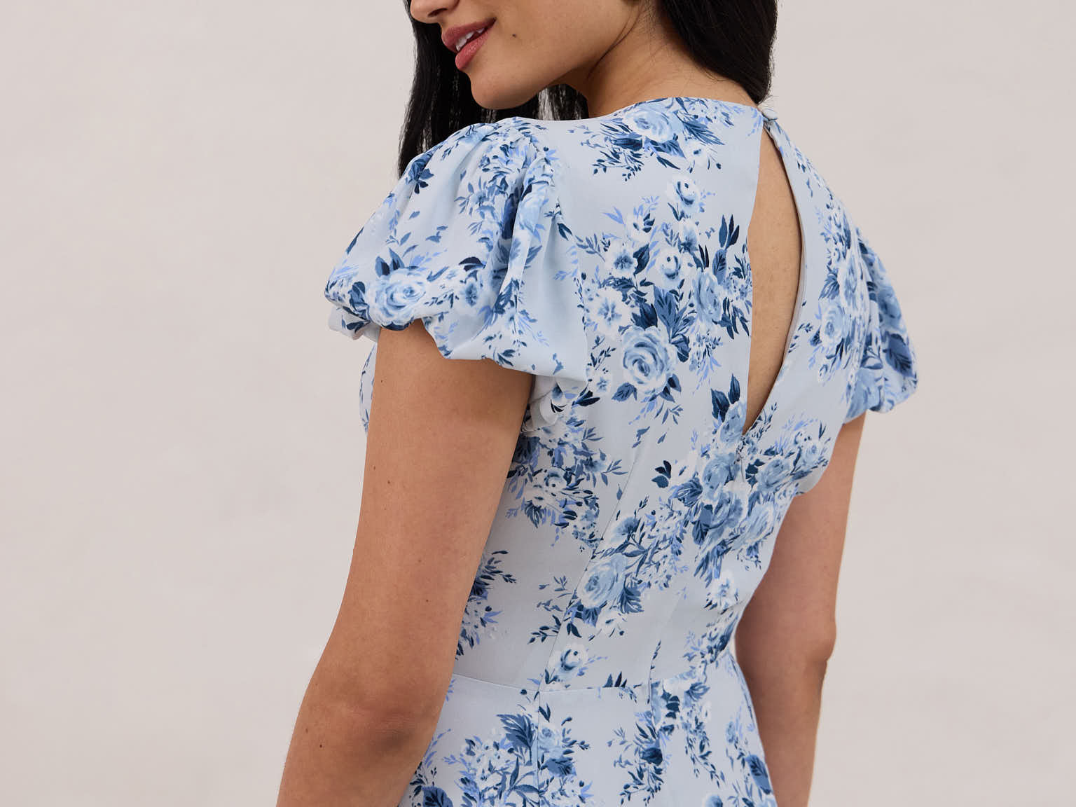 A woman is wearing the Beatrice Dress in Lila. This is a light blue dress with a beautiful blue floral print. The dress has a slightly puffed sleeve, and a full back with a keyhole detail. This is a bra-friendly dress. 