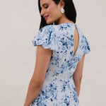 A woman is wearing the Beatrice Dress in Lila. This is a light blue dress with a beautiful blue floral print. The dress has a slightly puffed sleeve, and a full back with a keyhole detail. This is a bra-friendly dress. 