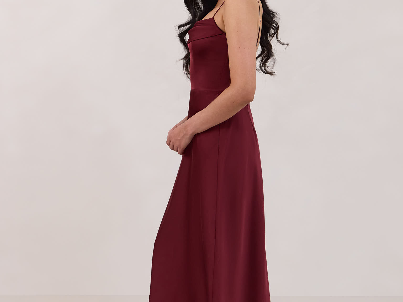 A woman is standing against a white backdrop wearing a burgundy, tea length satin bridesmaid dress. The dress has a boned bodice with a slight gathering at the bust, and a bias cut skirt that falls nicely over the hips. This is the Rosedale dress in merlot.