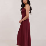 A woman is standing against a white backdrop wearing a burgundy, tea length satin bridesmaid dress. The dress has a boned bodice with a slight gathering at the bust, and a bias cut skirt that falls nicely over the hips. This is the Rosedale dress in merlot.