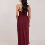 A woman is standing against a white backdrop facing the back wearing a burgundy, tea length satin bridesmaid dress. The dress has a boned bodice with a slight gathering at the bust, and a bias cut skirt that falls nicely over the hips. This is the Rosedale dress in merlot.
