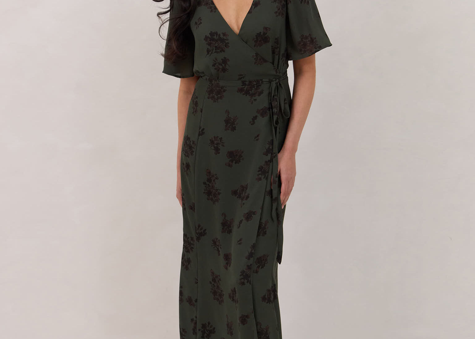 A woman is standing against a white backdrop wearing a dark green wrap dress with a subtle black floral print. The dress has a deep V neckline and loose sleeves. This is the Sherwood Dress in Esme.