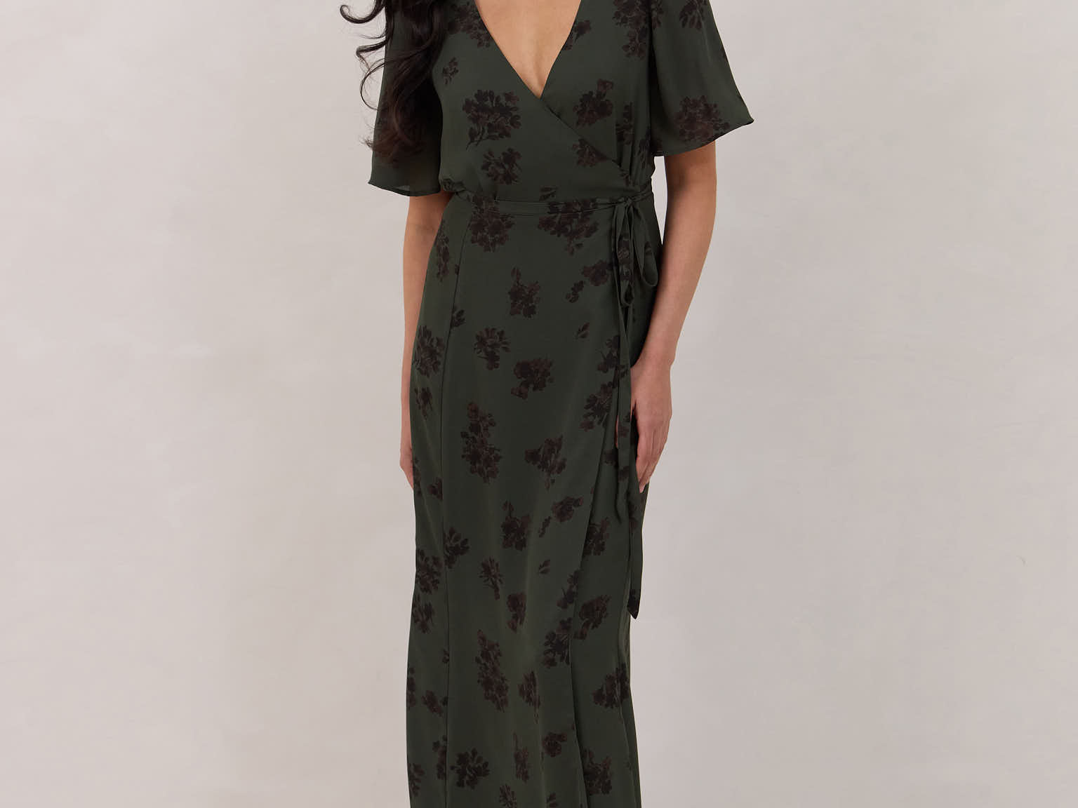 A woman is standing against a white backdrop wearing a dark green wrap dress with a subtle black floral print. The dress has a deep V neckline and loose sleeves. This is the Sherwood Dress in Esme.