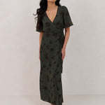 A woman is standing against a white backdrop wearing a dark green wrap dress with a subtle black floral print. The dress has a deep V neckline and loose sleeves. This is the Sherwood Dress in Esme.