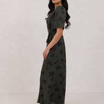 A woman is standing against a white backdrop facing the left is wearing a dark green wrap dress with a subtle black floral print. The dress has a deep V neckline and loose sleeves. This is the Sherwood Dress in Esme.