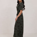 A woman is standing against a white backdrop facing the left is wearing a dark green wrap dress with a subtle black floral print. The dress has a deep V neckline and loose sleeves. This is the Sherwood Dress in Esme.