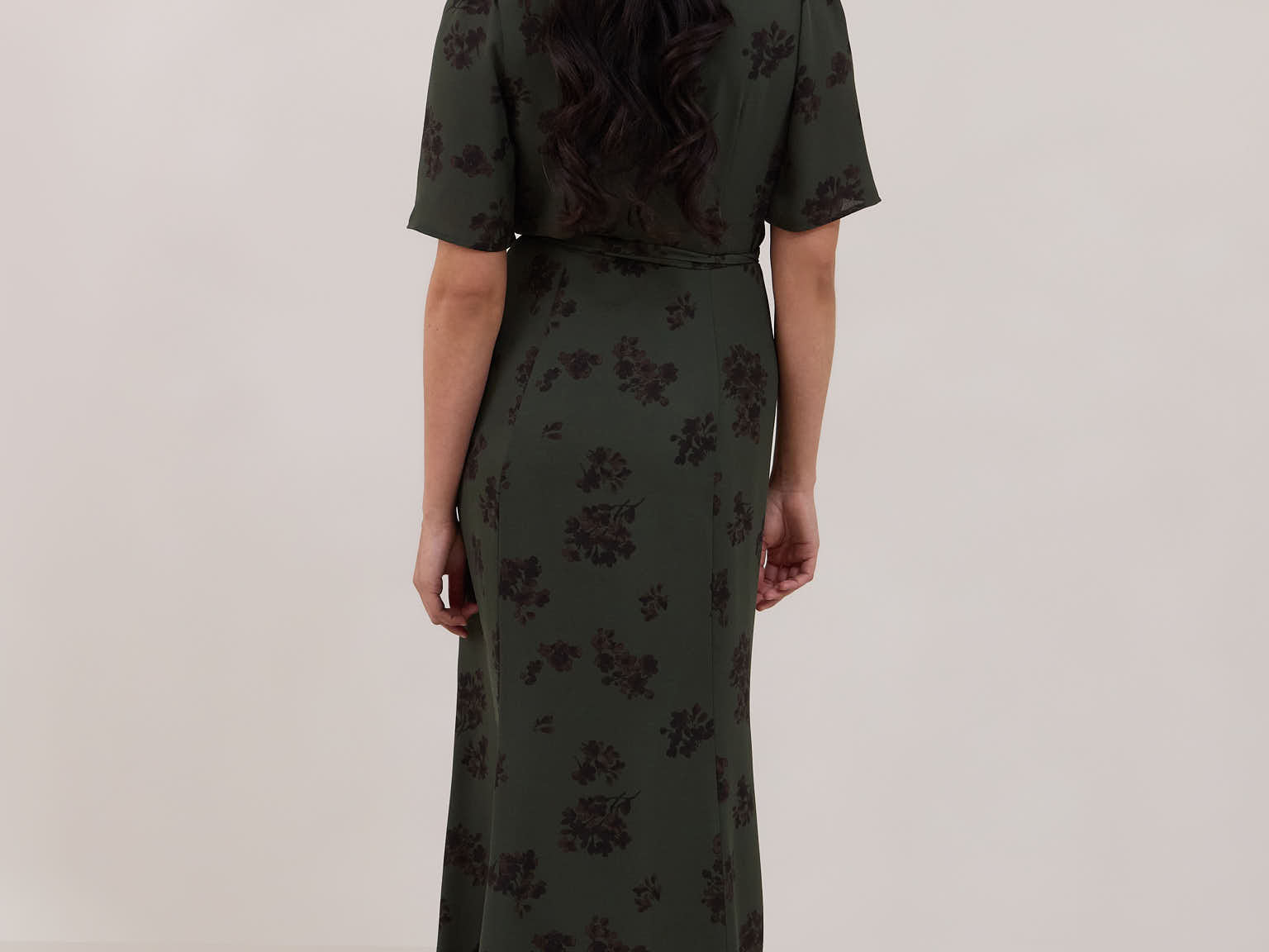 A woman is standing against a white backdrop facing the back is wearing a dark green wrap dress with a subtle black floral print. The dress has a deep V neckline and loose sleeves. This is the Sherwood Dress in Esme.
