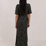 A woman is standing against a white backdrop facing the back is wearing a dark green wrap dress with a subtle black floral print. The dress has a deep V neckline and loose sleeves. This is the Sherwood Dress in Esme.