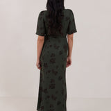 A woman is standing against a white backdrop facing the back is wearing a dark green wrap dress with a subtle black floral print. The dress has a deep V neckline and loose sleeves. This is the Sherwood Dress in Esme.