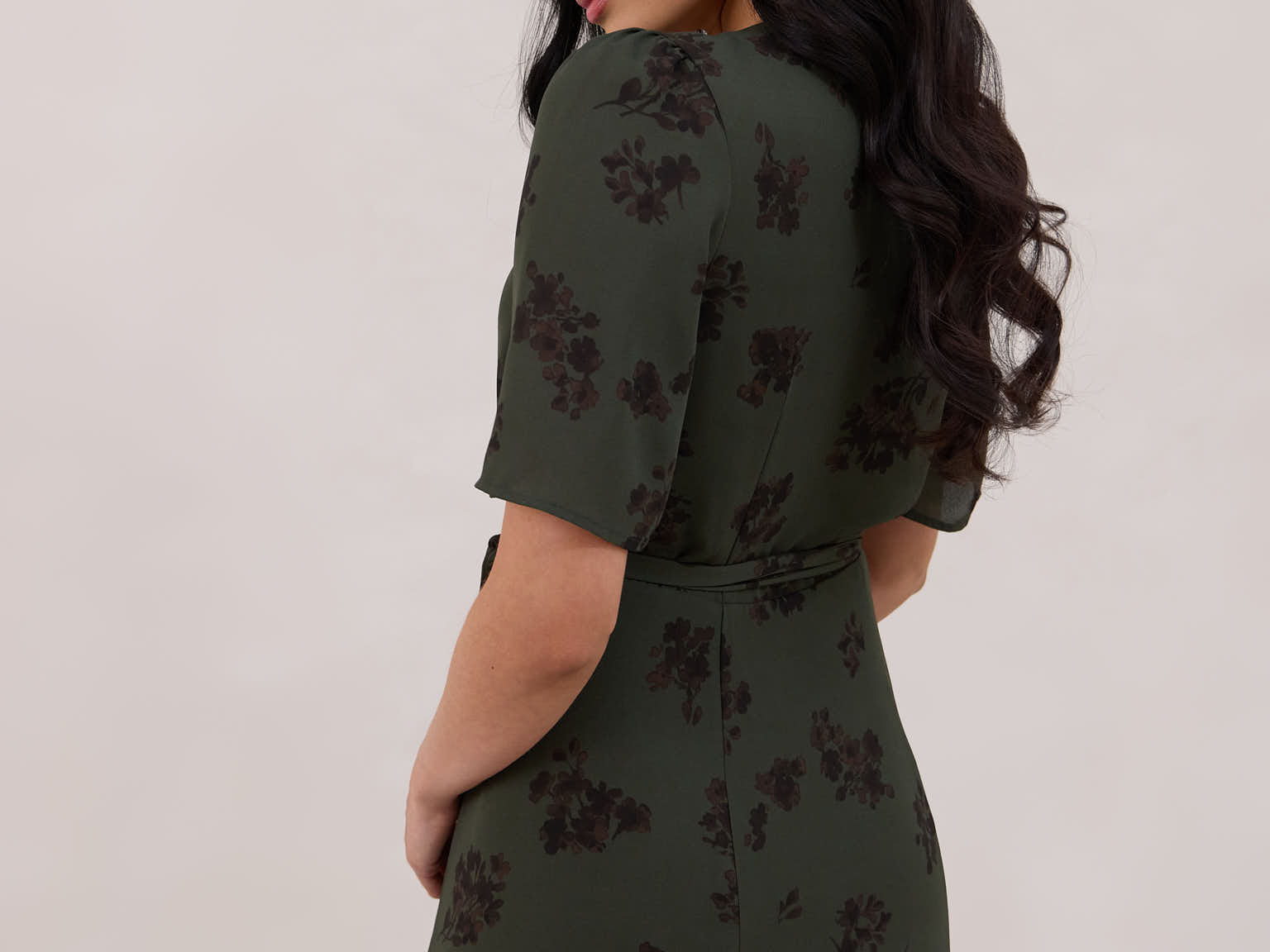 A woman is standing against a white backdrop facing the back is wearing a dark green wrap dress with a subtle black floral print. The dress has a deep V neckline and loose sleeves. This is the Sherwood Dress in Esme.