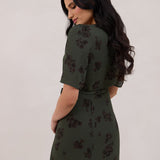A woman is standing against a white backdrop facing the back is wearing a dark green wrap dress with a subtle black floral print. The dress has a deep V neckline and loose sleeves. This is the Sherwood Dress in Esme.