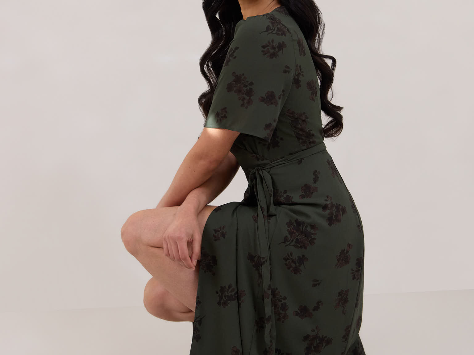 A woman against a white backdrop crouching down slightly is wearing a dark green wrap dress with a subtle black floral print. The dress has a deep V neckline and loose sleeves. This is the Sherwood Dress in Esme.