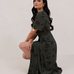 A woman against a white backdrop crouching down slightly is wearing a dark green wrap dress with a subtle black floral print. The dress has a deep V neckline and loose sleeves. This is the Sherwood Dress in Esme.