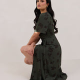 A woman against a white backdrop crouching down slightly is wearing a dark green wrap dress with a subtle black floral print. The dress has a deep V neckline and loose sleeves. This is the Sherwood Dress in Esme.