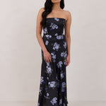 A woman is wearing a dark blue strapless dress with a light blue floral print. The dress is fitted in the bodice and hip then flows into a fuller skirt. The dress is tea-length. This is the Sterling Dress in Viola. 