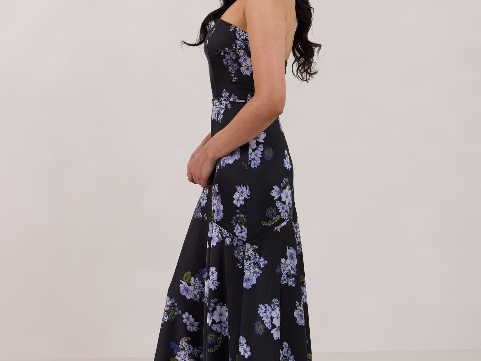 A woman is wearing a dark blue strapless dress with a light blue floral print. The dress is fitted in the bodice and hip then flows into a fuller skirt. The dress is tea-length. This is the Sterling Dress in Viola. 