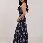 A woman is wearing a dark blue strapless dress with a light blue floral print. The dress is fitted in the bodice and hip then flows into a fuller skirt. The dress is tea-length. This is the Sterling Dress in Viola. 