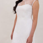 A woman stands against a white backdrop. She's wearing a delicate lace overlay dress on top of a simple white strapless mini bridal dress. This is the Winslow Overlay. 