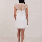 A woman stands against a white backdrop. She's wearing a delicate lace overlay dress on top of a simple white strapless mini bridal dress. This is the Winslow Overlay. 