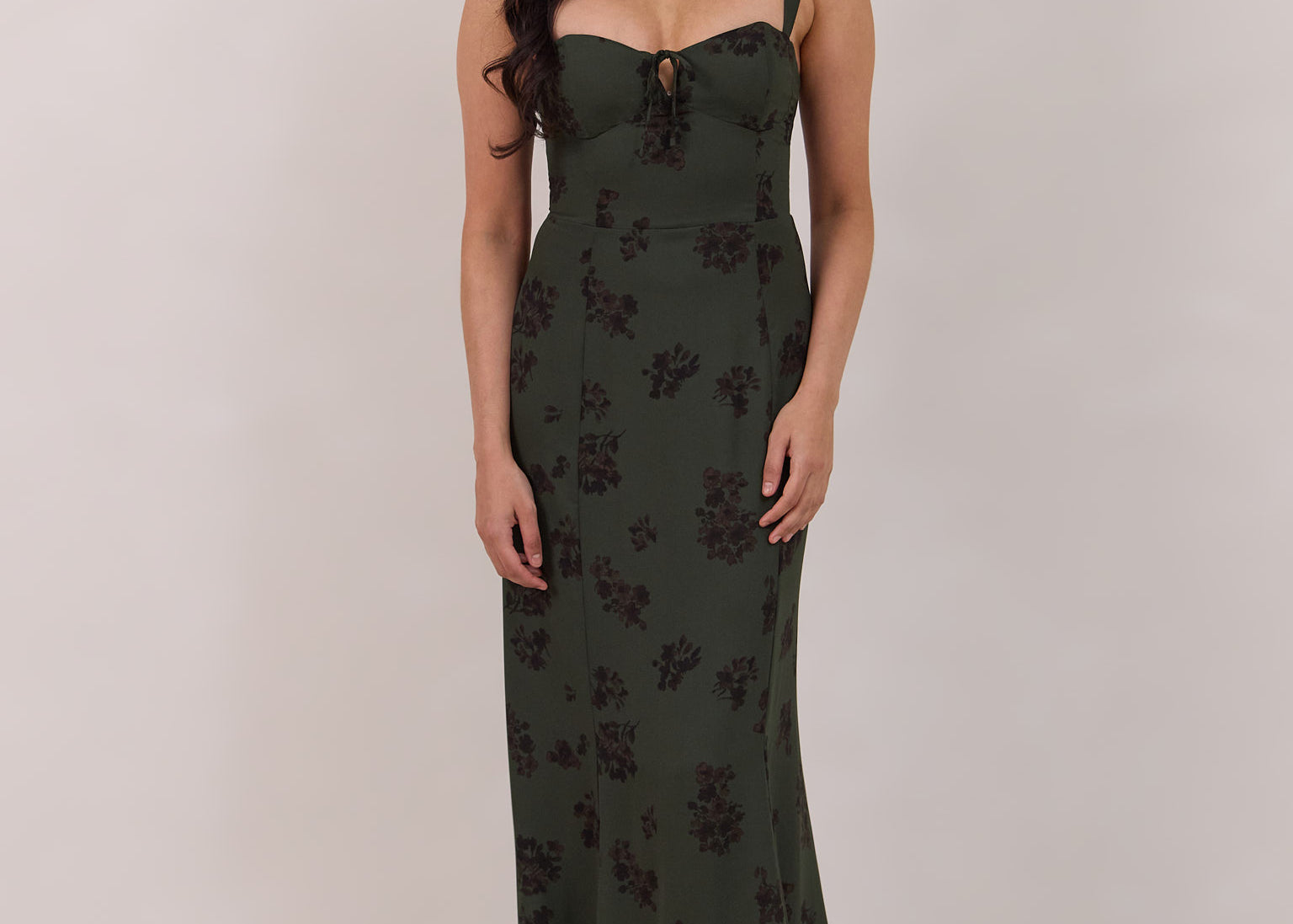A woman is standing infront of a floral display. She's wearing a dark green special occasion dress with a dark brown floral print. The dress s fitted in the bodice with a little keyhole detail and tie at the bust. The dress is tea-length and has shirring at the back for added comfort. This is the Decker dress in Esme.
