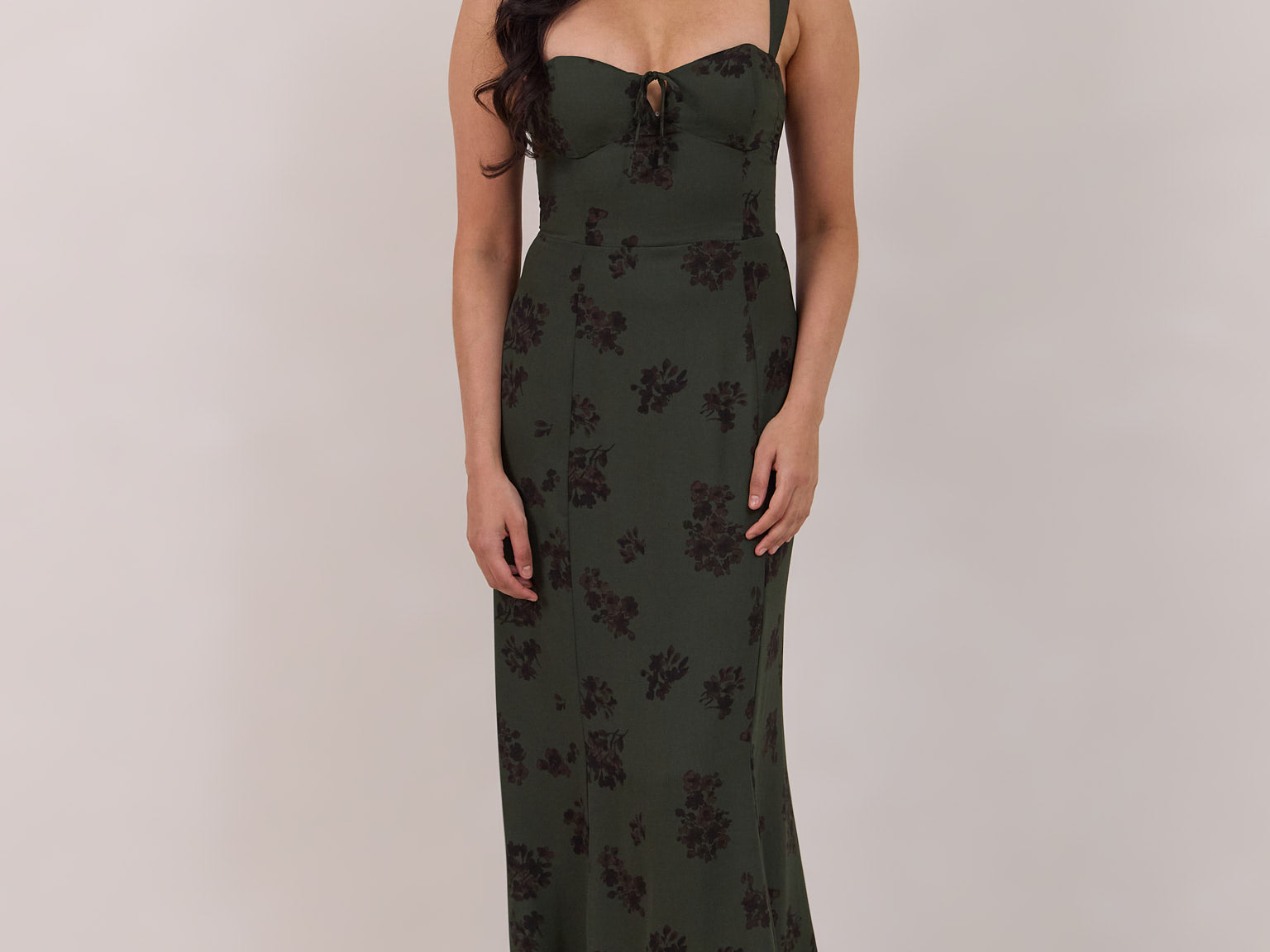 A woman is standing infront of a floral display. She's wearing a dark green special occasion dress with a dark brown floral print. The dress s fitted in the bodice with a little keyhole detail and tie at the bust. The dress is tea-length and has shirring at the back for added comfort. This is the Decker dress in Esme.