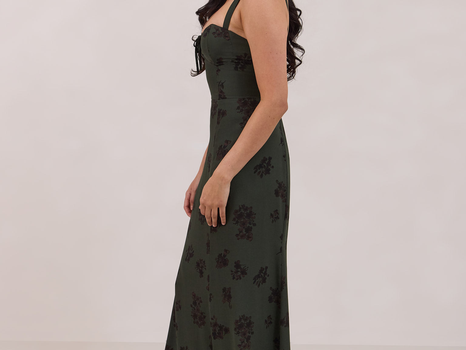 A woman is standing infront of a floral display. She's wearing a dark green special occasion dress with a dark brown floral print. The dress s fitted in the bodice with a little keyhole detail and tie at the bust. The dress is tea-length and has shirring at the back for added comfort. This is the Decker dress in Esme.