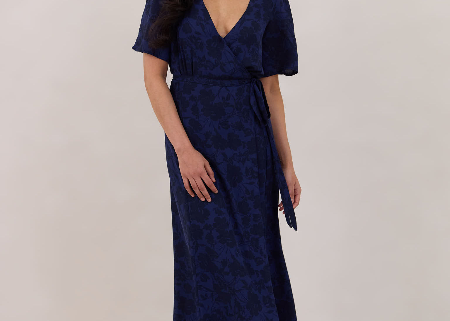 A woman is standing against a white backdrop wearing a navy blue wrap dress with a subtle black floral print. The dress has a deep V neckline and loose sleeves. This is they Sherwood Dress in Twyla.