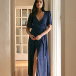 A woman is standing in a hallway wearing a navy blue wrap dress with a subtle black floral print. The dress has a deep V neckline and a loose sleeve. This is the Sherwood Dress in Twyla. 