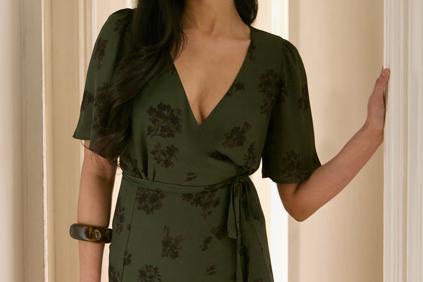 A woman is standing in a hallway wearing a dark green wrap dress with a subtle black floral print. The dress has a deep V neckline and loose sleeves. This is the Sherwood Dress in Esme.