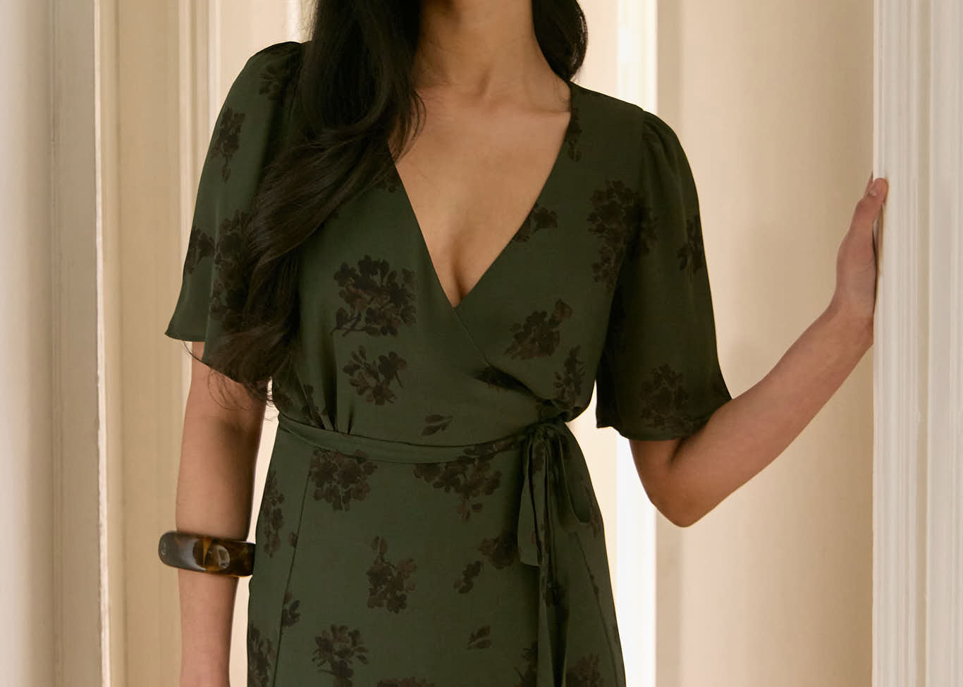 A woman is standing in a hallway wearing a dark green wrap dress with a subtle black floral print. The dress has a deep V neckline and loose sleeves. This is the Sherwood Dress in Esme.