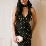 A woman stands against a white backdrop wearing a black and white polka dot dress. The dress has a halter neckline with an optional scarf detail. She's holding a white clutch. This is the Archer Dress in Midnight Dot.