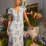 A woman is standing infront of a floral display wearing the Beatrice Dress in Lila. This is a light blue dress with a beautiful blue floral print. The dress has a slightly puffed sleeve, and a full back with a keyhole detail. The front has a deep-V cut with a delicate tie at the bust and another keyhole detail down to the waistline. This is a bra-friendly dress that is ankle length. 