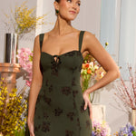 A woman is standing infront of a floral display. She's wearing a dark green special occasion dress with a dark brown floral print. The dress s fitted in the bodice with a little keyhole detail and tie at the bust. The dress is tea-length. This is the Decker dress in Esme.