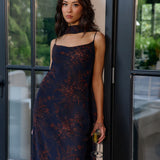 Woman in a dark floral dress holding a glass, standing in front of a glass door.